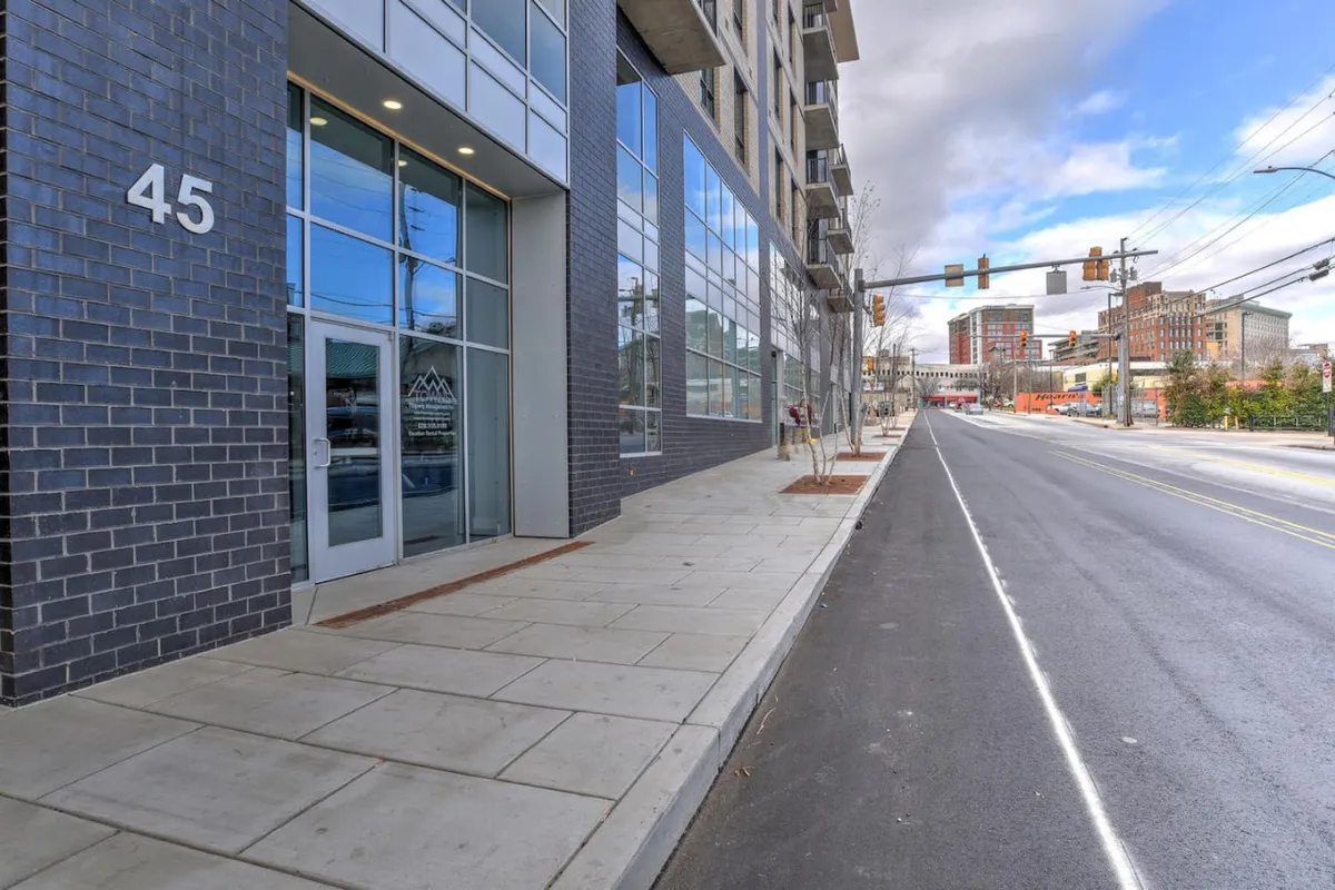 A lively exterior shot of the condo building captures its modern facade with large glass windows fronting a bustling, walkable street. People walk along the sidewalk, enjoying coffee at a nearby cafe, while the vibrant city atmosphere is highlighted by the soft glow of sunset. The number 45 is clearly visible above the entrance, situating the property in the heart of the action.