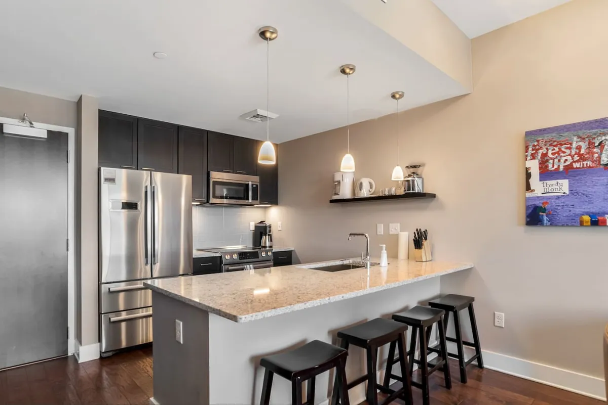 The image displays a contemporary kitchen with a sleek island, stainless steel appliances, and modern bar stools. Artwork on the wall adds a touch of personality, while the overall layout is open and designed for both cooking and socializing. The space is well-lit and inviting, making it ideal for culinary endeavors and gatherings.