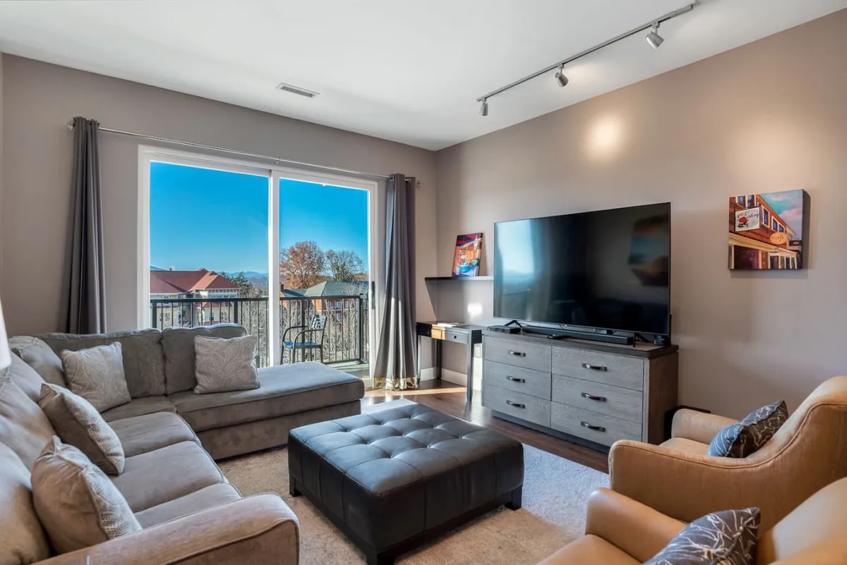 A modern, cozy living area featuring a large window that fills the room with natural light. The space includes a comfortable sofa, a media console with a TV, and a stylish armchair, creating a welcoming environment ideal for relaxing, watching movies, or enjoying heartfelt conversations. The view from the window hints at an urban setting, connecting the indoors to the city outside.