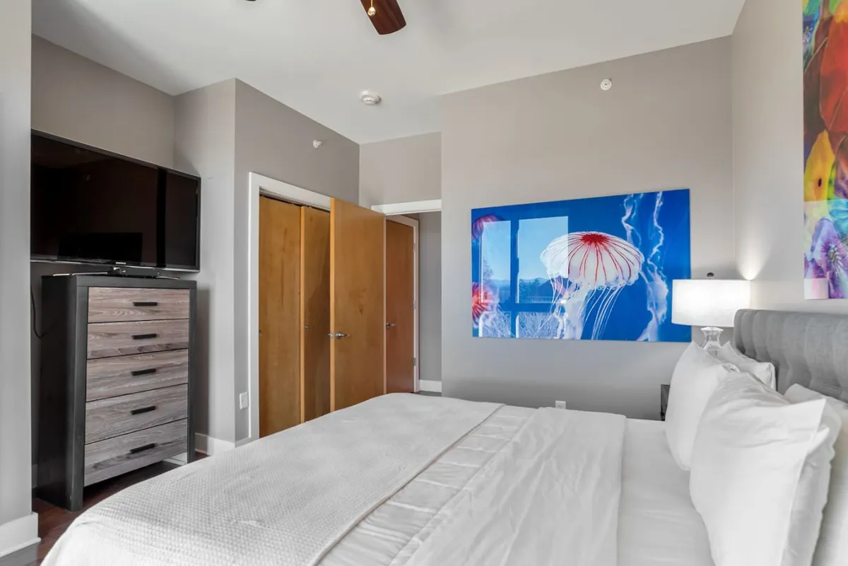 A tranquil king bedroom featuring a plush bed with crisp white linens, accented by colorful jellyfish artwork above the headboard. A television sits atop a sleek dresser, offering entertainment options. The room is flooded with natural light, creating a serene retreat ideal for restful nights or unwinding after a busy day.