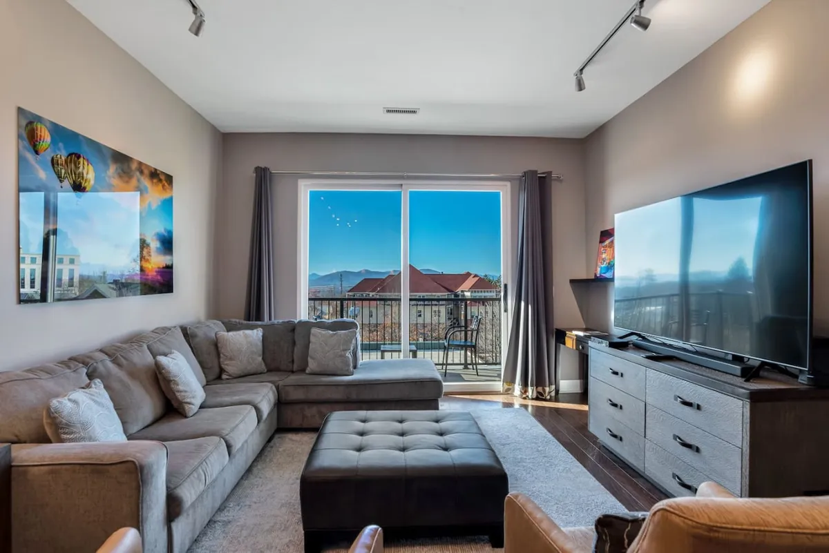 A modern living room featuring a large sectional sofa, flat-screen TV, sliding glass doors to a balcony, and colorful wall art. The space feels spacious and open, with direct access to the outdoors—a nod to easy movement within the home.