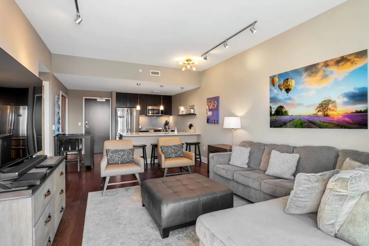 An inviting, open-plan interior that seamlessly connects the living area with the kitchen and dining space. The scene highlights a comfortable sofa, eye-catching artwork, a kitchen island, and dining chairs, all set within a warm, welcoming atmosphere ideal for both entertaining and casual relaxation.