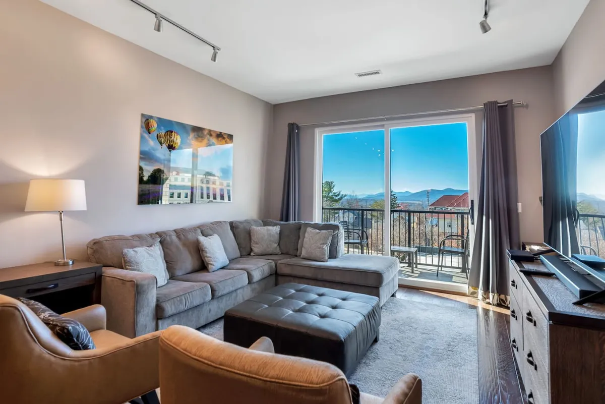 The image showcases a stylish living area with a plush sofa, striking local artwork, and sliding doors opening onto a balcony with stunning sunrise views over Asheville. The scene radiates comfort and warmth, inviting guests to relax indoors or step outside to take in the cityscape and mountain backdrop.