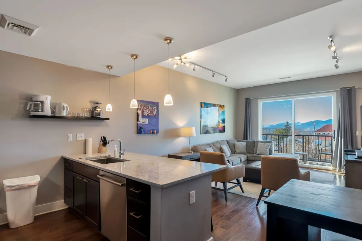 A contemporary interior featuring a kitchen area adjacent to a cozy sofa seating arrangement, with a view extending out to a balcony. The space is awash with natural light, creating an inviting atmosphere for cooking, relaxing, or enjoying sunrise views.